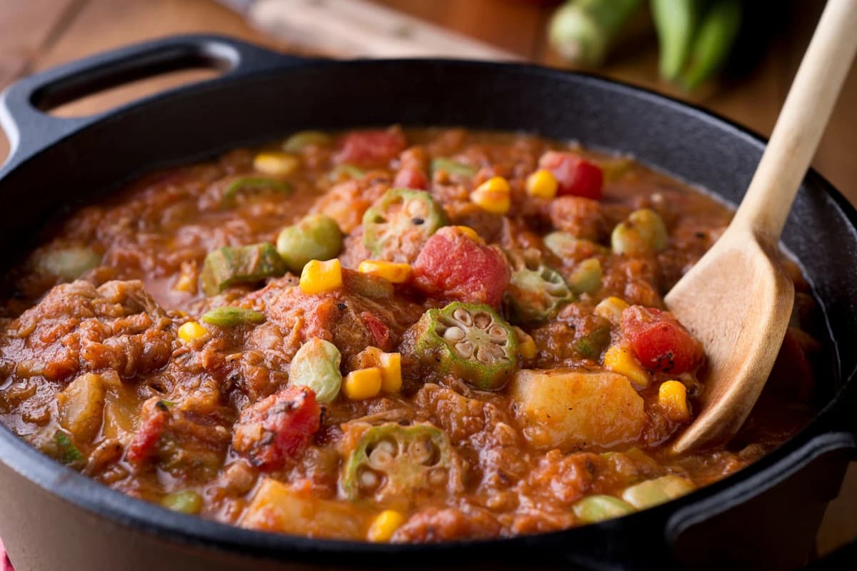 Hearty tomato-based stew with okra, corn, and potatoes simmering in a cast-iron pot.