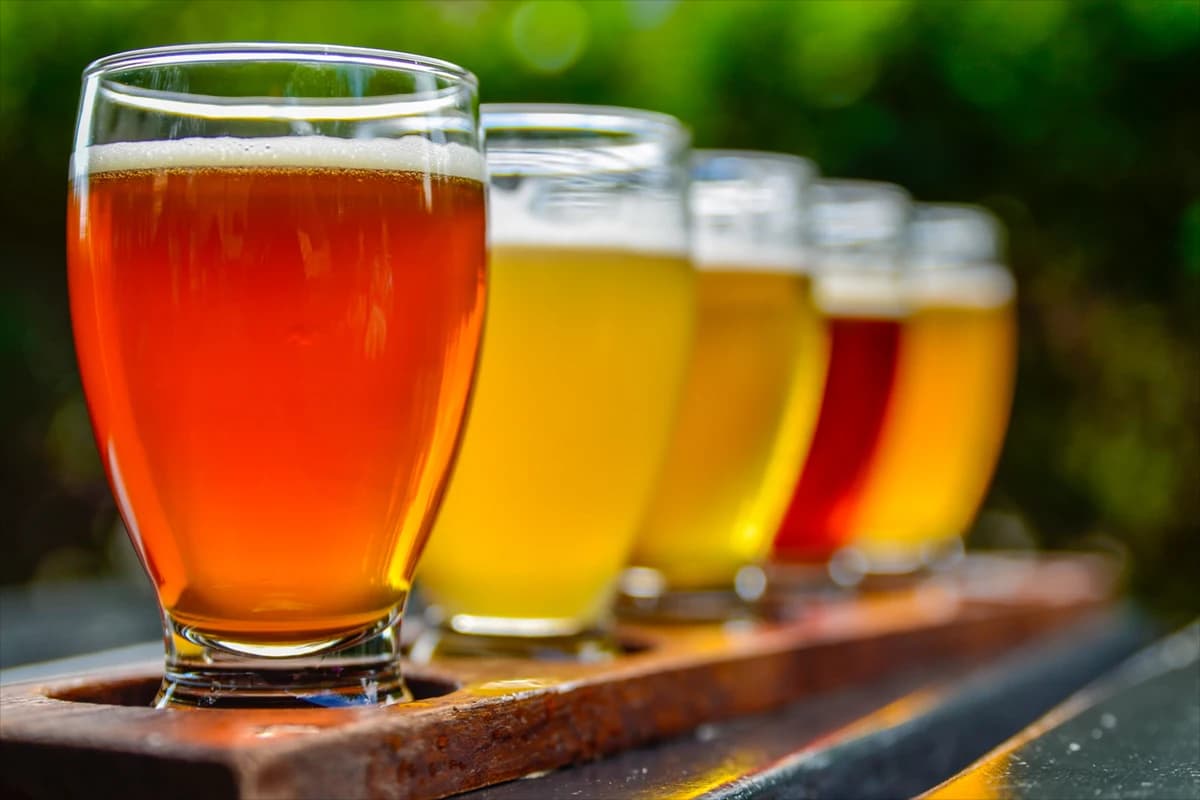 Flight of craft beers in clear pint glasses on a wooden paddle outdoors.