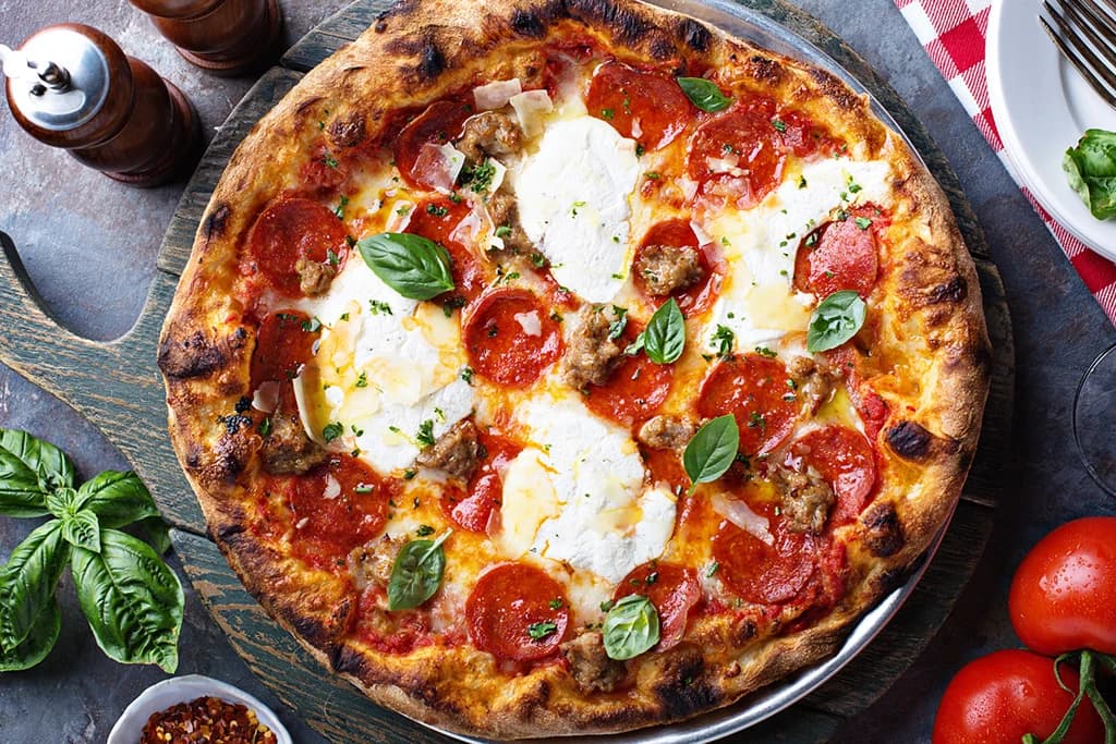 Wood-fired pepperoni and sausage pizza with ricotta dollops, fresh basil, and a blistered crust on a rustic board.